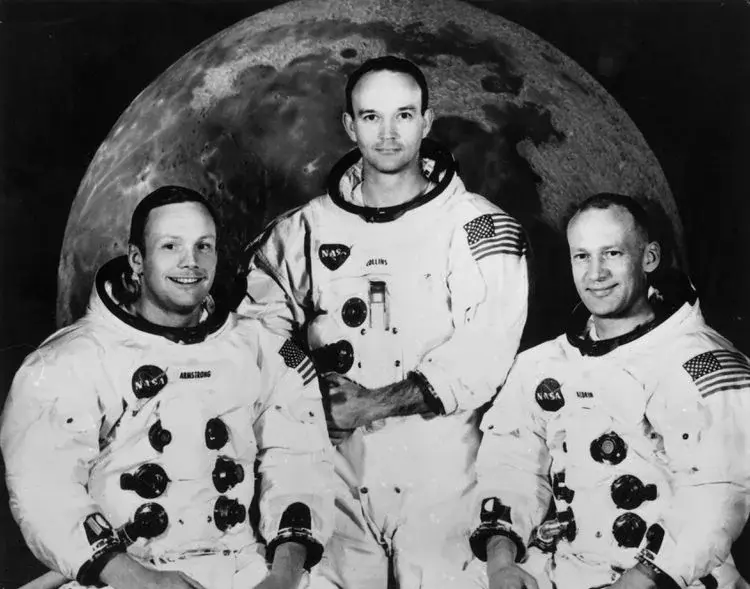 Ασπρόμαυρο πορτρέτο του πληρώματος του Apollo 11, Armstrong, Collins και Aldrin, με στολές μπροστά σε φωτογραφία της Σελήνης